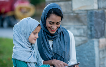 A Muslim woman and her daughter are outdoors. They are wearing casual clothes and head scarves. They are sitting in front of a stone wall. They are watching something funny on a tablet computer.